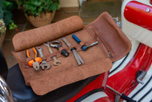 Open brown leather motorcycle tool roll laid out on the seat of a red motorcycle, displaying various tools including a wrench, pliers, and a screwdriver
