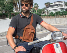 Man wearing a black polo shirt and blue jeans, sitting on a red scooter, with a brown leather crossbody bag featuring detailed stitching and multiple pockets. Handcrafted men's brown leather crossbody bag with detailed stitching, adjustable buckle and strap, and multiple pockets. Perfect for stylish and functional everyday use.