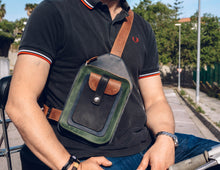 Man wearing a black polo shirt and blue jeans, with a handcrafted men's leather crossbody bag featuring green, brown, blue, and black leather, detailed stitching, and multiple pockets, sitting on a scooter.