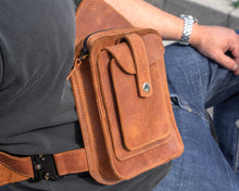 Close-up of a handcrafted brown leather men's crossbody bag with detailed stitching and multiple pockets, worn over a black polo shirt and blue jeans