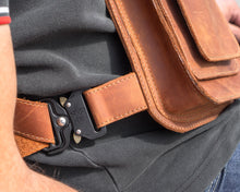Close-up of a brown leather crossbody bag with detailed stitching, showing the buckle and strap, worn over a dark green polo shirt.