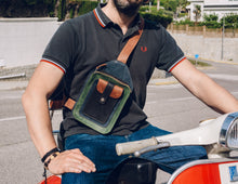 Man wearing a black polo shirt and blue jeans, with a handcrafted men's leather crossbody bag featuring green, brown, blue and black leather, detailed stitching, and multiple pockets. Featuring a sturdy buckle and adjustable strap