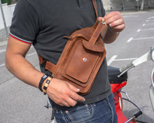 Close-up of a man wearing a black polo shirt and blue jeans, holding a handcrafted brown leather men's crossbody bag with detailed stitching and multiple pockets, standing next to a red scooter