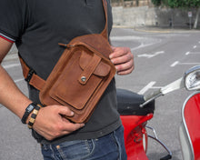 Close-up of a man wearing a black polo shirt and blue jeans, holding a brown leather crossbody bag with detailed stitching and multiple pockets, standing next to a red scooter