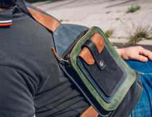 Man wearing a dark green polo shirt and blue jeans, holding a handcrafted men's leather crossbody bag with green, brown, blue, and black leather, detailed stitching, and multiple pockets.