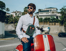 Man wearing a white shirt and blue jeans, with a handcrafted men's leather crossbody bag featuring green, brown, blue, and black leather, detailed stitching, and multiple pockets, sitting on a red scooter in a residential neighborhood