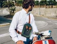 Man wearing a white shirt and blue jeans, with a handcrafted men's leather crossbody bag featuring green, brown, blue, and black leather, detailed stitching, and multiple pockets, sitting on a red scooter and looking to the side.