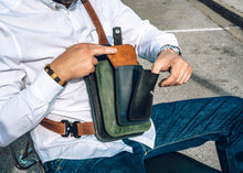 Man wearing a white shirt and blue jeans, showing a handcrafted men's leather crossbody bag with green, brown, blue, and black leather, detailed stitching, multiple pockets, and an adjustable strap with a sturdy buckle.