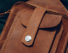 Close-up of a brown leather crossbody bag with detailed stitching and a snap button closure.