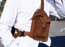 Close-up of a man's brown leather crossbody bag featuring detailed stitching and multiple pockets, worn over a white shirt and blue jeans.