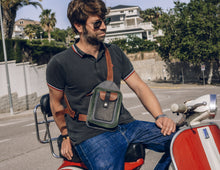 Man wearing a black polo shirt and blue jeans, with a handcrafted men's leather crossbody bag featuring green, brown, blue, and black leather, detailed stitching, multiple pockets, a sturdy buckle, and an adjustable strap, sitting on a red scooter