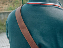 lose-up of a brown leather strap from a crossbody bag over the shoulder of a person wearing a dark green polo shirt with red and white trim."