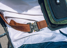 Close-up of a handcrafted men's leather crossbody bag with green, brown, and black leather, detailed stitching, and an adjustable strap with a sturdy buckle, worn over a white shirt.