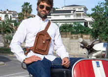 Close-up of a man sitting on a red scooter, wearing a white shirt and blue jeans, with a brown leather crossbody bag featuring detailed stitching and multiple pockets.