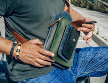 Man wearing a dark green polo shirt and blue jeans, holding a handcrafted men's leather crossbody bag with green, brown, blue, and black leather, showing multiple pockets and detailed stitching.