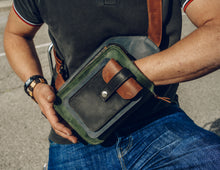 Man wearing a dark green polo shirt and blue jeans, holding a handcrafted men's leather crossbody bag with green, brown, blue, and black leather, detailed stitching, and multiple pockets