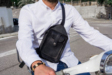 Man wearing a white shirt and blue jeans, riding a scooter, showcasing a men's black leather crossbody bag with detailed stitching and multiple pockets.