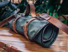 Rolled waxed canvas and leather knife bag with adjustable carrying straps on wooden table