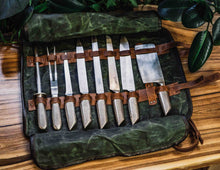 Handcrafted waxed canvas and leather knife roll bag open on wooden table, holding professional chef knives, carving fork, and sharpening steel in leather loops