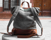 Rear view of handcrafted grey waxed canvas backpack with padded straps and brown leather base, artisan roll-top rucksack on stone bench