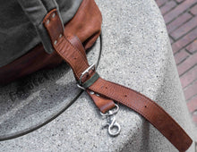 Close-up of brown leather backpack strap with silver buckle and hook clasp, attached to grey waxed canvas and leather base