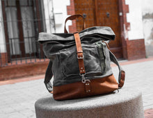 Handcrafted grey waxed canvas and brown leather roll-top backpack with front pocket, styled on urban stone bench near wooden door