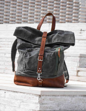 Stylish grey waxed canvas backpack with brown leather strap and base, roll-top design displayed on white brick steps