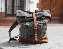 Handcrafted grey waxed canvas backpack with brown leather straps and base, vintage roll-top design displayed on stone bench