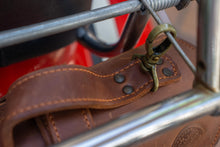 Close-up of a fastening clip on a brown leather motorcycle tool roll, securely attached to the chrome frame of a motorcycle, highlighting the sturdy hardware.