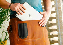 Handmade leather apron with a large leather pocket and brass details, showcasing a person placing a tablet in the pocket. Ideal for professionals like chefs, bartenders, or baristas looking for a custom leather apron for their daily work