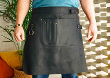 A black leather apron designed for professionals in the kitchen or barista space, with a customized strap and heavy-duty brass hardware. Perfect for chefs and bartenders
