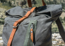Close-up of grey waxed canvas backpack with brown leather strap and zipper pull, showcasing roll-top and front pocket stitchin