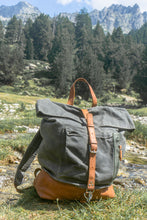 Grey waxed canvas roll-top backpack with brown leather base and strap, standing on grassy mountain trail near stream
