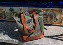 Artisan tote bag made of waxed canvas with green trim and brown leather base, placed on a stone bench with colorful Spanish ceramic tile backdrop