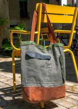 Large waxed canvas and leather tote bag with front pocket, hanging on a yellow chair outdoors, showcasing green, navy, and brown color palette