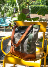 Handcrafted waxed canvas tote bag with brown leather bottom and handles, displayed on a yellow metal chair in a sunny garden setting