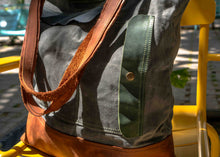Detail shot of durable waxed canvas tote bag featuring green leather trim and raw brown leather handles, on a yellow chair in natural light