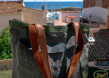 Close-up of leather and canvas tote bag showing detailed stitching and green upper trim, with coastal Mediterranean town in background