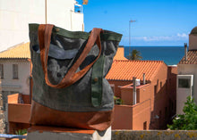 Stylish canvas tote bag with green, brown, and navy panels and leather straps, displayed against a scenic seaside rooftop view