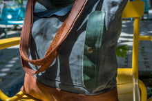 Detail shot of durable waxed canvas tote bag featuring green leather trim and raw brown leather handles, on a yellow chair in natural light