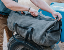 Close-up of a person zipping up a large, rugged gray waxed canvas duffel bag with black leather straps, placed on industrial equipment. The person is wearing a blue shirt and a wooden bracelet.