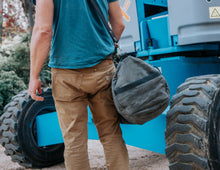 Man carrying a large, rugged gray waxed canvas duffel bag with black leather straps and a shoulder strap, wearing a blue shirt and beige pants, walking next to industrial equipment.