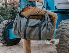Man climbing industrial equipment, carrying a large, rugged gray waxed canvas duffel bag with black leather straps and a shoulder strap, wearing a blue shirt, beige pants, and white sneakers.