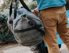 Man sitting on industrial equipment, unzipping a large, rugged gray waxed canvas duffel bag with black leather straps and a shoulder strap, wearing a blue shirt, beige pants, and white sneakers.