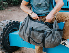 Man carrying a large, rugged gray waxed canvas duffel bag with black leather straps and a shoulder strap, wearing a blue shirt, beige pants, and white sneakers, standing next to industrial equipment.