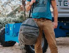 Man carrying a large, rugget gray waxed canvas duffel bag with black leather straps and a shoulder strap, wearing a blue shirt and beige pants, standing next to industrial equipment