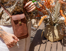 Alt Text: Woman holding a handmade leather phone crossbody bag. Description: A woman wearing a floral blouse and light-colored pants is holding a handmade leather phone crossbody bag. The bag is brown with a front flap secured by a brass button and features a long strap worn across the body. The background includes a wooden table and decorative flowers.