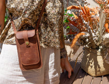 Alt Text: Woman showcasing a handmade leather phone crossbody bag. Description: A woman wearing a floral blouse and light-colored pants is showcasing a handmade leather phone crossbody bag. The bag is brown with a front flap secured by a brass button and features a long strap worn across the body. The background includes a wooden table and orange flowers.