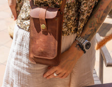 Alt Text: Handmade leather phone crossbody bag with open flap. Description: A handmade leather phone crossbody bag with its front flap open, showing the inside. The bag is brown with a long strap. The background includes a wooden table and decorative flowers.