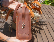 Alt Text: Back view of a handmade leather phone crossbody bag. Description: The back view of a handmade leather phone crossbody bag, showing a small external pocket with a snap closure. The bag is brown with a long strap. The background includes part of a wooden table and some decorative flowers.